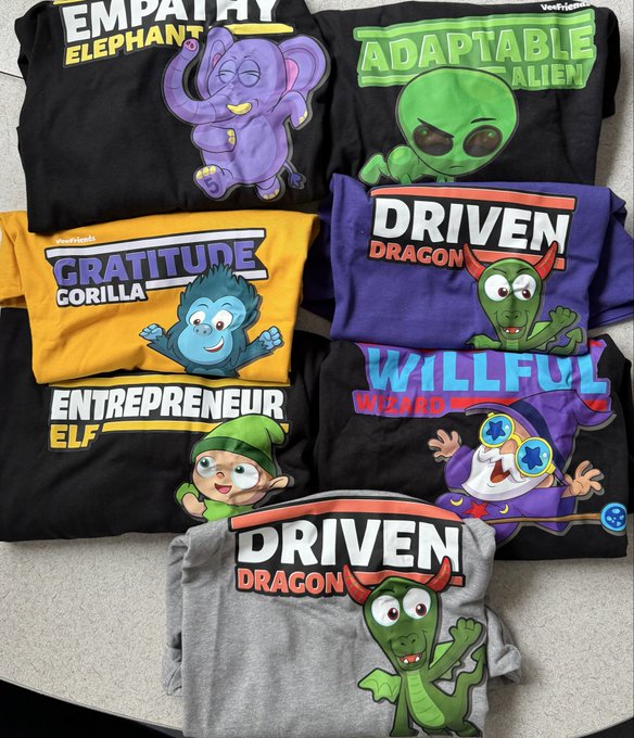 A collection of folded VeeFriends t-shirts is displayed on the floor, featuring characters paired with positive traits like "Empathy Elephant," "Adaptable Alien," "Gratitude Gorilla," "Driven Dragon," "Entrepreneur Elf," and "Willful Wizard," connecting to the post about receiving fresh VeeFriends swag at home after a flight cancellation.