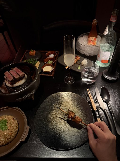 An intimate fine-dining setting featuring wagyu beef on a hot stone, a small dish of fried rice, condiments, skewers, sake, and sparkling water on a dark table.