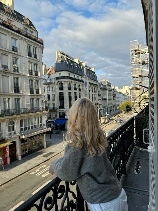 A blonde woman leans over a Parisian balcony railing, overlooking a classic Haussmann-style street with elegant cream-colored buildings and a stop sign visible below. The image complements the "gm who's actually locking in today?" post by pairing a dreamy, leisurely city view with a call to hustle, creating a relatable contrast between escapism and productivity.