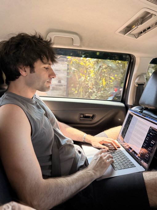 The image shows a man sitting inside a car, using his laptop while wearing a grey tank top. The scene includes a window next to him that offers a view of trees outside. There are also other people visible in the background who appear to be engaged in their own activities or conversations.