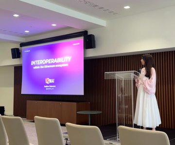 A woman in a white dress speaking at a podium during a presentation, with a large screen behind her displaying 'INTEROPERABILITY within the blockchain ecosystem' alongside the IQAI logo.