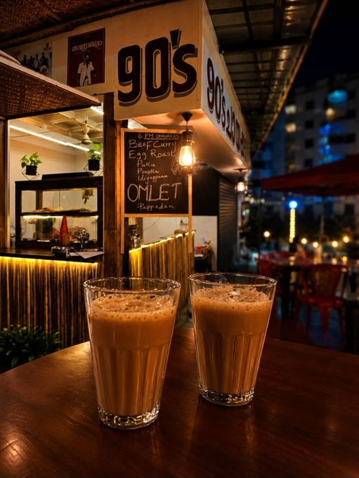 Two glasses of milky tea sit on a wooden table at a street-side tea stall named "90's," with a chalkboard menu listing items like "Beef Curry," "Egg Roast," and "Omlet Pappadam."