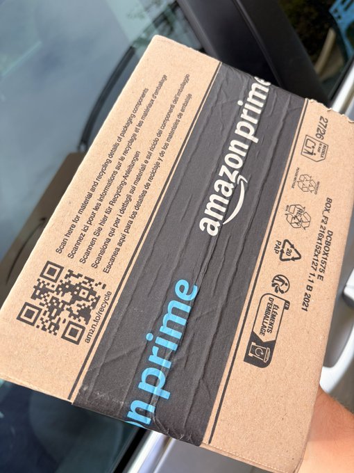 A hand holds a brown Amazon Prime cardboard shipping box displaying a QR code and recycling information inside a vehicle.