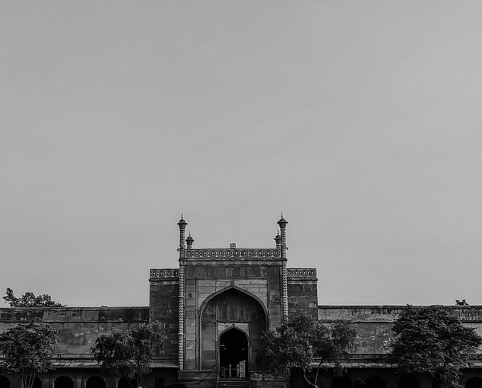 A black-and-white photograph shows a historic stone gateway with two minarets, centered between long walls with trees in the foreground under an overcast sky.