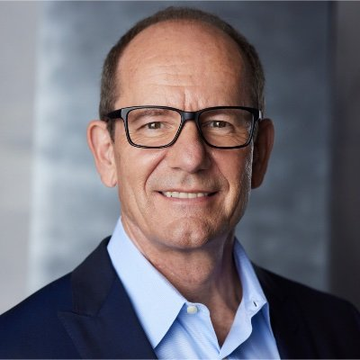 A professional headshot of an older man with glasses, wearing a dark blazer over a light blue shirt, smiling at the camera against a blurred background.