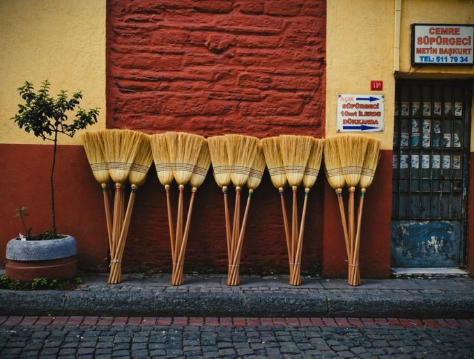 Five traditional straw brooms with wooden handles lean against a red and yellow stone wall outside a shop with Turkish signage.