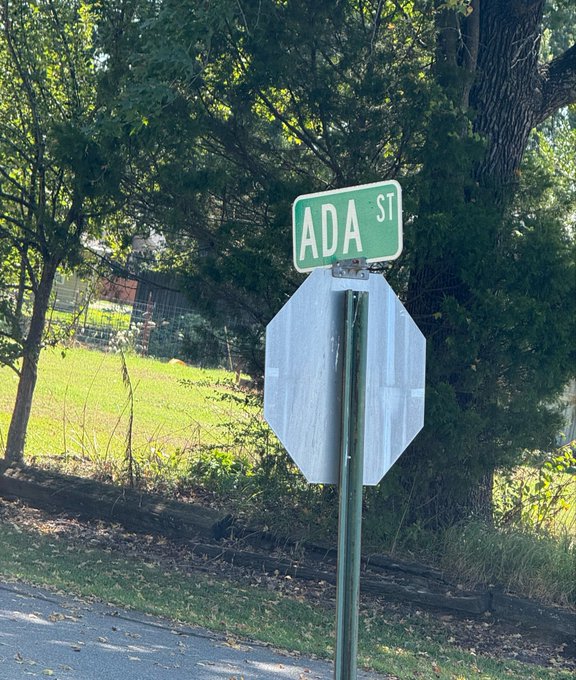 A street sign reading 'ADA ST' mounted above a stop sign, with the back facing the camera, set against a backdrop of trees and a grassy yard.
