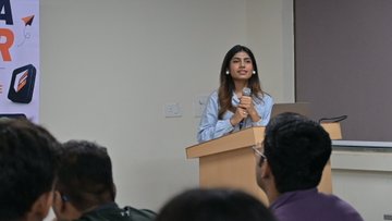A woman speaks at a podium to an audience in what appears to be a classroom or conference setting.