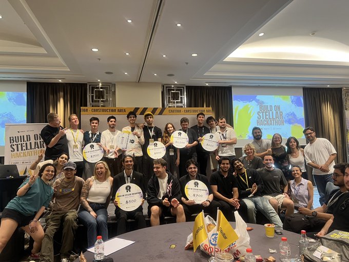 The image shows a group of people posing for a photo at an Istanbul Hackathon event. There are 13 individuals in total, with some standing and others sitting around a table. They all have papers or cards in their hands, which could be related to the hackathon. The room has a large screen on one side and a dining table nearby.

The post mentions that this is the second edition of the Istanbul Hackathon, and it highlights the achievements of the local community. It also mentions the winners of the event, who have been recognized for their hard work and dedication to the hackathon. The image captures the spirit of collaboration and innovation in the context of a successful hackathon.