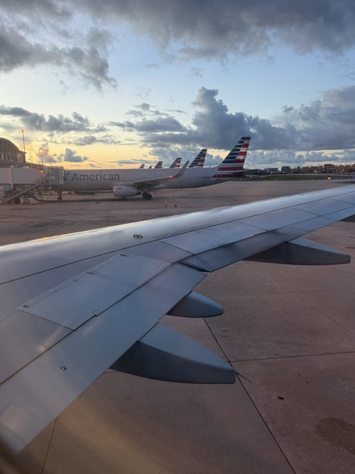 An American Airlines plane is visible through the window with its wing in the foreground at sunset, indicating the traveler is en route to Colombia for the Primavera Eterna art residency.