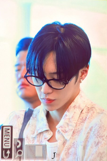 A person with short dark hair wearing black-framed glasses and a white patterned shirt looks downward, appearing to be in a candid moment likely at a fan event or public appearance. A "MENU" interface with camera settings icons is visible in the bottom left corner, suggesting the photo was taken in an app with editing features, while the "J" watermark and hashtags (#ALPHADRIVEONE, #ALD1, #ARNO) indicate the image is connected to a member of the K-pop group ALPHA DRIVE ONE.
