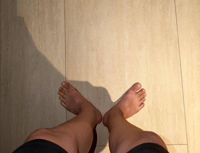 A pair of bare feet stand on a sunlit tiled floor, casting a shadow, jokingly visualizing the "full send" partnership announcement with the $TOESCOIN meme, as if the person stepped off to seal the deal.