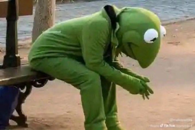 A person in a Kermit the Frog costume sits hunched over on a bench by the water, head bowed in a pose often used to depict sadness or disappointment.
