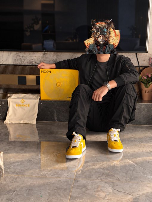 A person wearing a striking cyberpunk-style cat/wolf mask and bright yellow-and-white sneakers sits on a marble floor next to a yellow "MOON" box and a beige "BINANCE" tote bag. The image ties into the post's review of Binance's German trainer sneakers, with the yellow shoes matching the MOON branding in the photo.