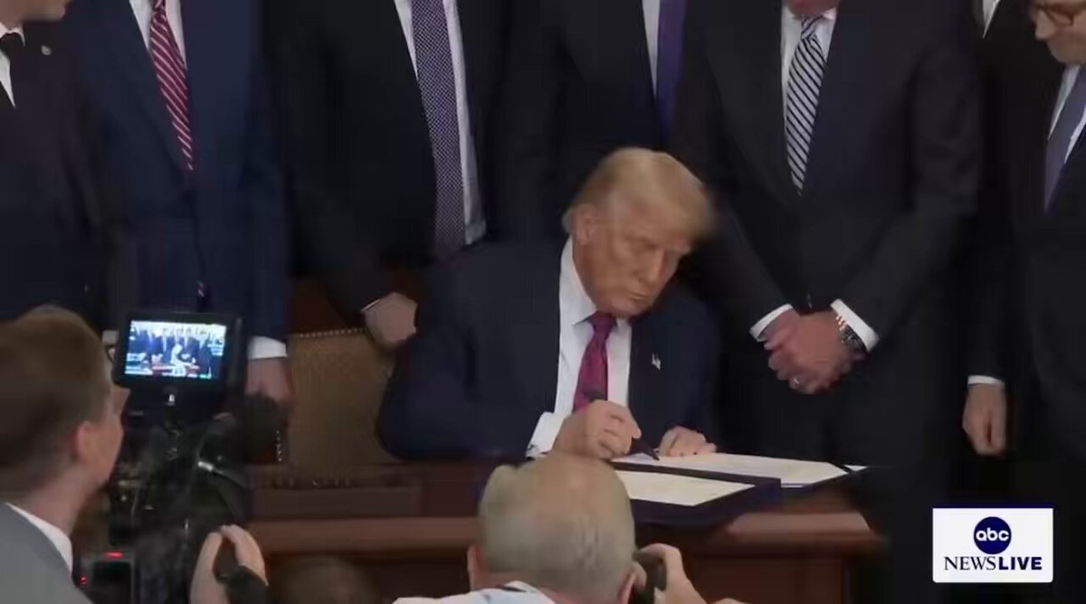 The image shows a man sitting at a desk signing a document while surrounded by other people who are taking pictures of him with cameras. The scene appears to be from a news broadcast, as indicated by the presence of a TV screen in the background. The focus on the man signing the document and the camera angles suggest that this is an important event or moment being covered by the news organization.