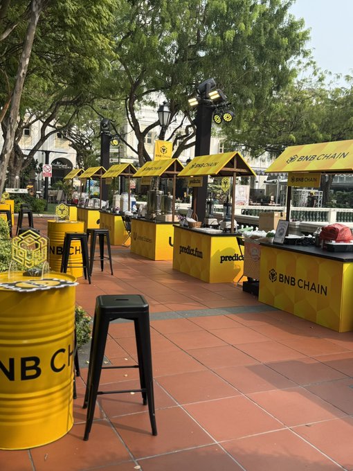 An outdoor crypto/Web3 event booth featuring yellow BNB CHAIN branded counters and stools, including a 'predicta.fm' stand and yellow barrel seating, set on a tiled plaza with trees.