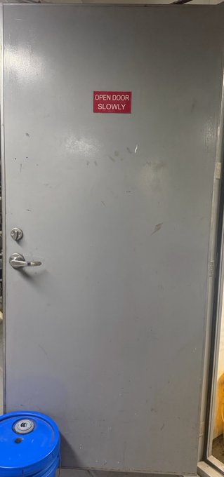 A gray metal door with a silver lever handle and a red sign reading 'OPEN DOOR SLOWLY' mounted near the top, with a blue barrel at the bottom left.