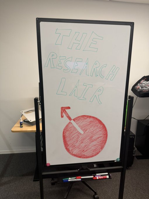 A whiteboard drawing labeled 'THE RESEARCH LAIR' featuring a red circle with an arrow, with markers on the tray below.