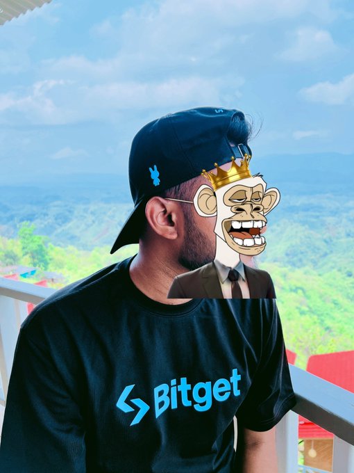 A photograph of a man wearing a dark blue 'Bitget' t-shirt and a backwards navy cap, with a cartoon monkey wearing a crown superimposed over his face, standing on a balcony with mountains in the background.