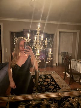 A mirror selfie of a woman with long blonde hair in a black dress, taken in an elegant room featuring a chandelier and a patterned rug.