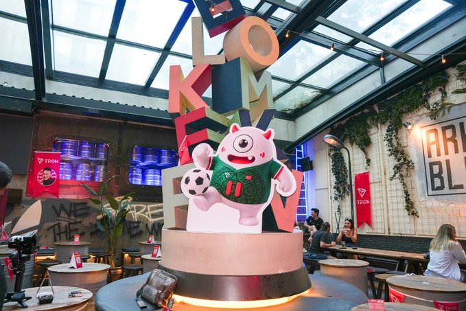 A pink mascot holding a soccer ball stands on a pedestal in front of large "TRON" letters at an indoor event venue, with visible "TRON" branding on banners and a sign reading "WE ARE HERE." The setup reflects the post's praise for a great TRON-themed gathering, capturing the celebratory atmosphere the user attributes to the project and its community event.
