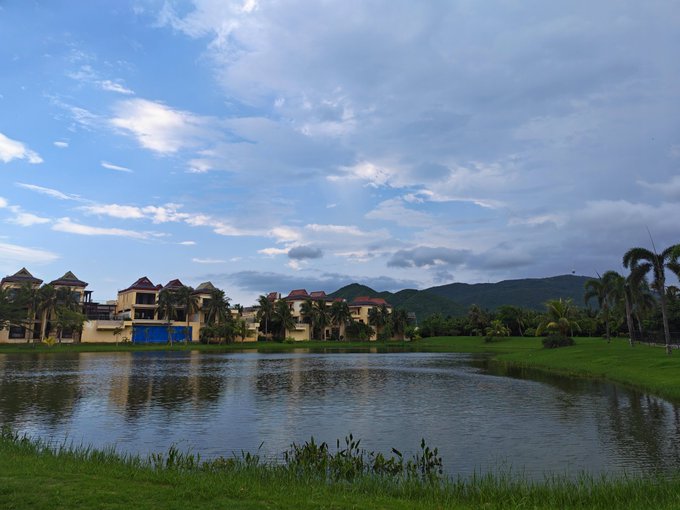 A tranquil landscape showing a pond or lake with buildings and palm trees along the far shore, set against rolling green hills and a partly cloudy blue sky.