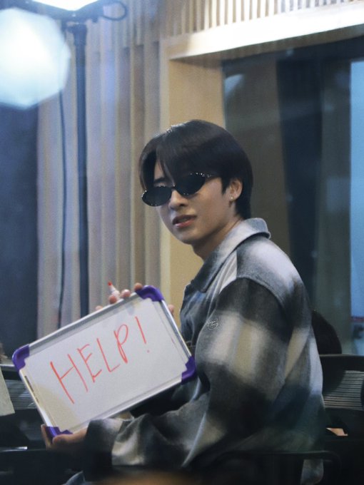 A young man with dark hair and black sunglasses holds up a small whiteboard with 'HELP!' written in red marker.