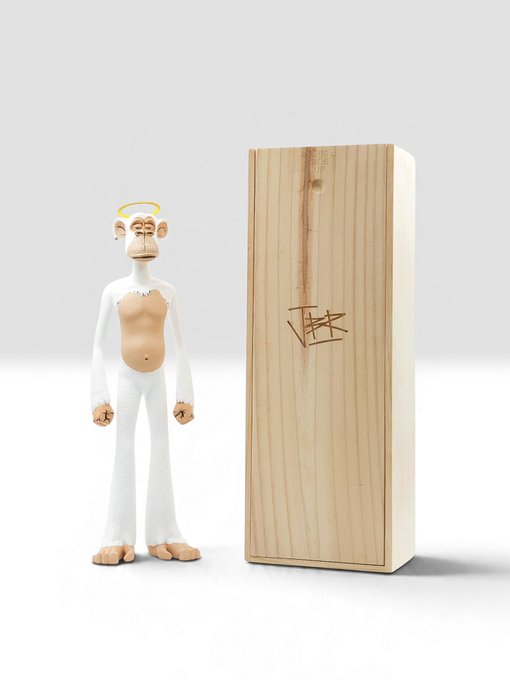 A small designer vinyl figure of a white, monkey-like character with an exposed tan torso and a yellow halo stands next to its wooden display box signed by the artist.