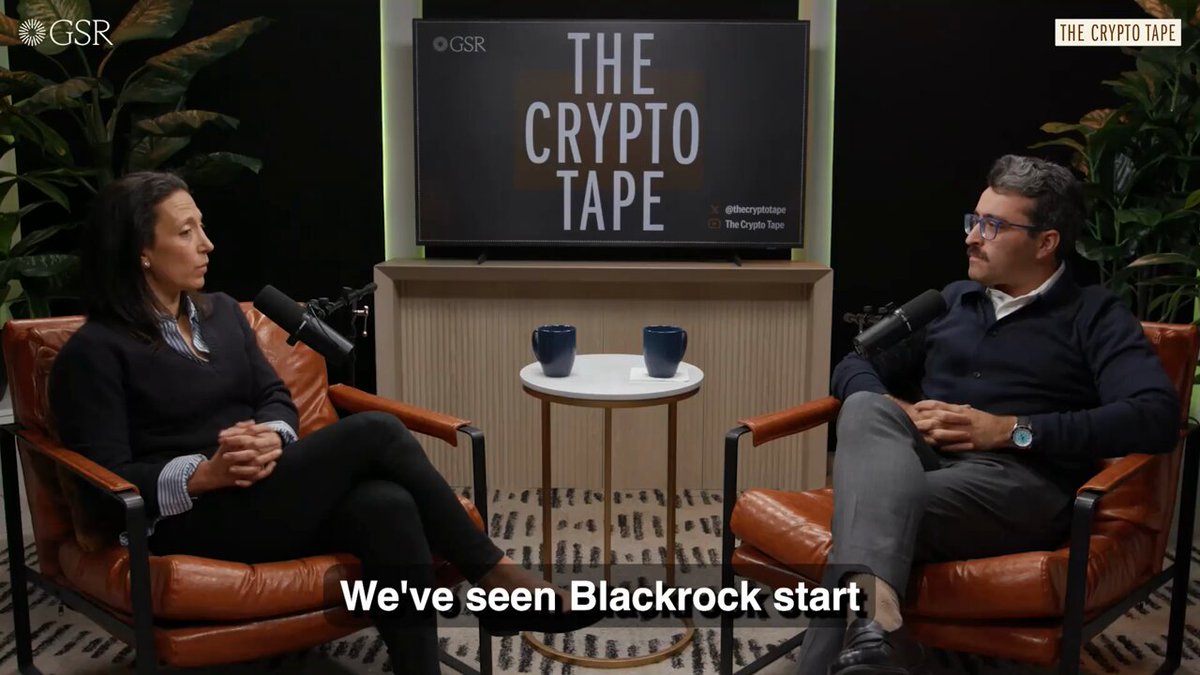 The image shows a man and woman sitting in chairs, talking about cryptocurrency. The background features a TV screen displaying "The Crypto Tape" logo. There are two cups placed between them, suggesting they might be enjoying some refreshments during their conversation.