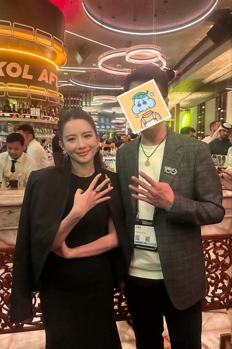 The image features a man and woman posing together, with the woman holding up her face over her shoulder as if she has a mask or costume on. The man is wearing a suit and tie, which adds an element of formality to their pose. They are standing in front of a bar, suggesting that they might be attending a social event or gathering.