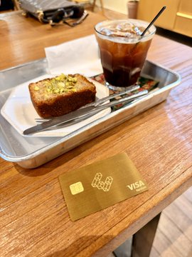 A slice of avocado toast and an iced coffee sit on a wooden table next to a gold Visa card embossed with "EtherFi," suggesting a casual café visit where the crypto payment card is being used to purchase the meal.