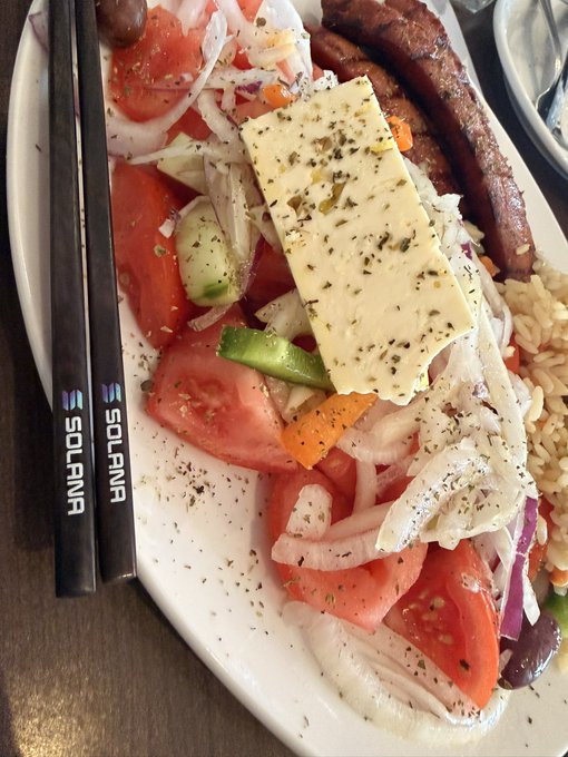 A Greek salad topped with a generous slab of feta cheese is served alongside grilled sausages and rice, with two black chopsticks branded "SOLANA" resting on the plate. The post highlights a creative (and self-admittedly silly) way to showcase the Solana-branded chopsticks by pairing them with a meal, turning an ordinary lunch into a fun crypto-themed photo opportunity.