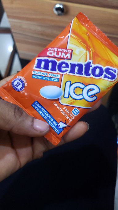 A hand holds a packet of Mentos Ice sugar-free chewing gum with xylitol in the orange/mint flavor.