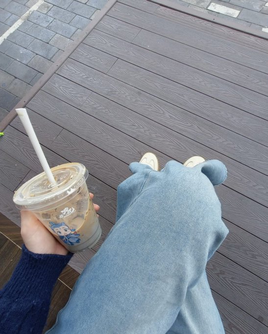 A first-person view looking down at a person's hand holding a plastic cup with a straw containing an iced coffee drink, alongside their legs clad in light blue jeans and white sneakers on a wooden deck.