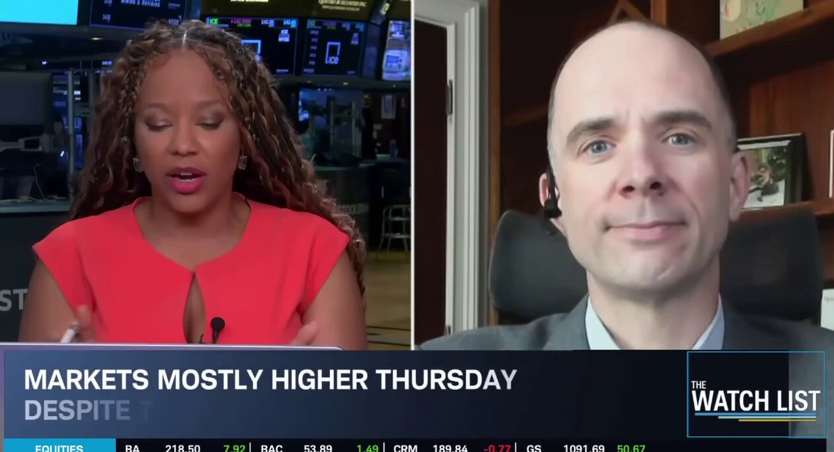 In the image, there are two people sitting behind computer screens with market graphs displayed during their conversation. They appear to be discussing the topic of Bitcoin's value. The person at the left is wearing a red shirt and holding a microphone. At the right side, the other person can be seen looking towards them. The backdrop features an article about bitcoin selling for Saylor Capital Markets more than 22 times higher on Thursday compared to its last price. The discussion revolves around this information while the two individuals focus their attention on the market graphs displayed in front of them.