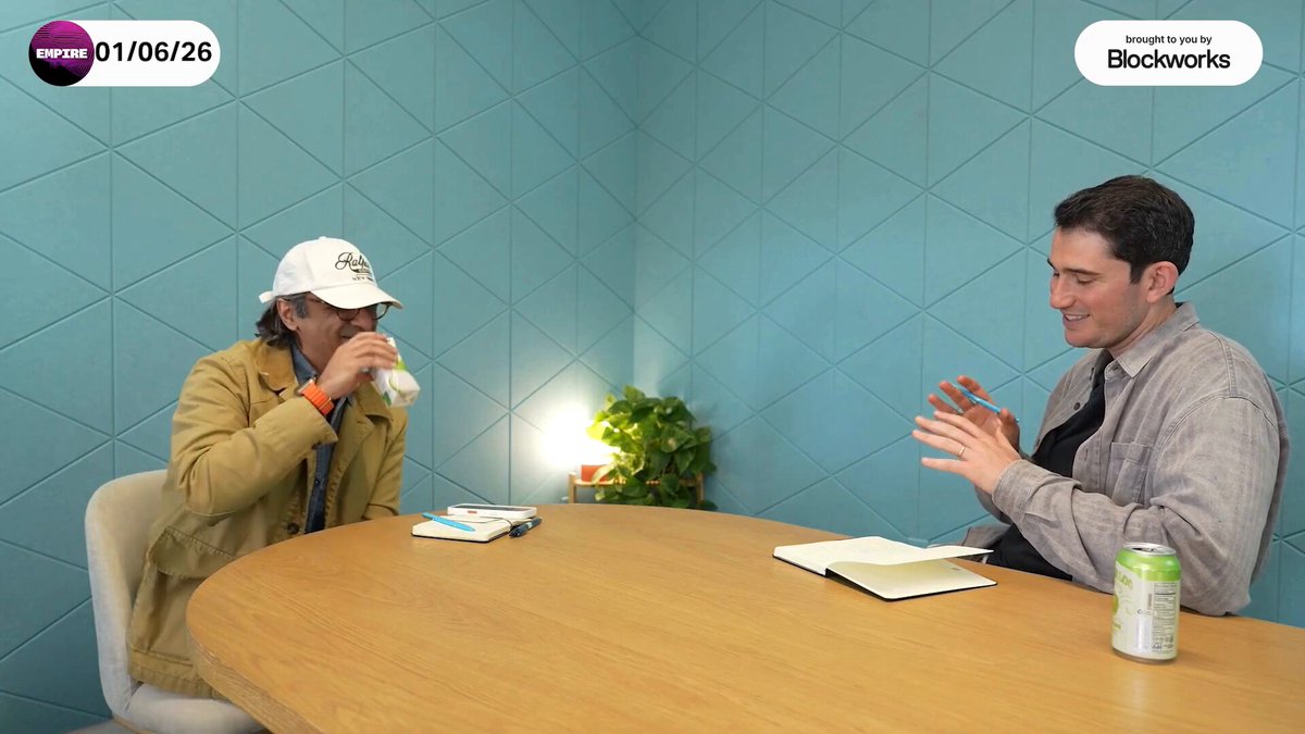 The image shows two men sitting at a table with their hands on their laps, engaged in conversation. One man has a drink in his hand while they talk. The background features blue walls that create an atmosphere for their discussion. A plant can be seen near them, adding some greenery to the scene. The post about Ethereum and the US weaponizing the dollar is related to the image as it discusses how Bitcoin and other cryptocurrencies are being used to combat the US dollar's dominance in the global economy.