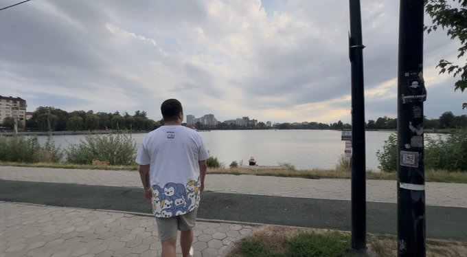 A man wearing a white t-shirt with blue cartoon characters on the bottom stands on a paved walkway looking out over a lake with a city skyline in the distance under a cloudy sky.