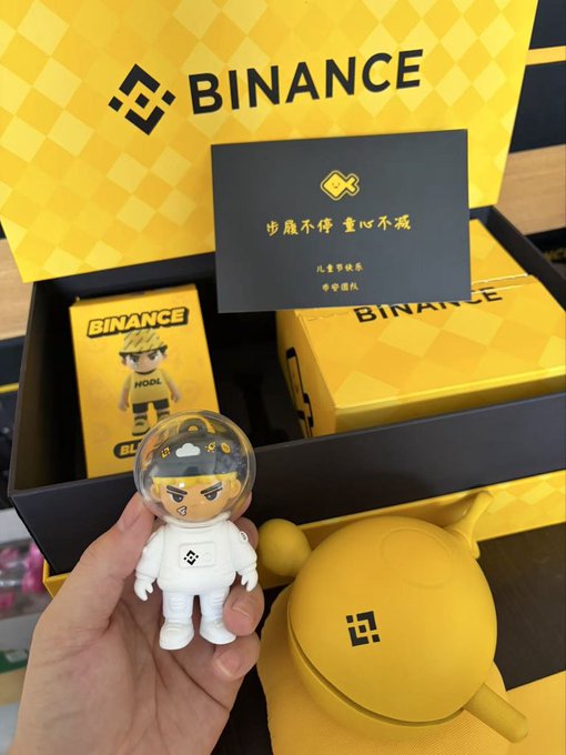 The image features a person holding up a small toy astronaut figurine, which appears to be yellow with a black helmet. The toy is placed next to a box containing more of these figurines, as well as other items such as a yellow ball and a card. The scene suggests that the person might have received or purchased these toys for themselves or someone else, possibly as a gift or for personal collection.