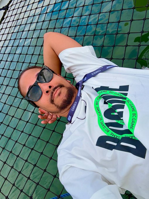 A relaxed man wearing sunglasses lies against a net with his hands behind his head, sporting a white "BC.GAME" t-shirt and a "BC.GAME" lanyard, with "GM" (Good Morning) greeting his followers.