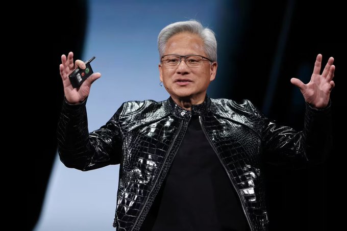 The image features a man wearing a black leather jacket, standing on stage with his arms outstretched as if he's giving a speech or presentation. The background of the image is dark, which contrasts with the bright light shining down on him from above. There are no other people in the scene, and the focus is solely on the man and his actions.