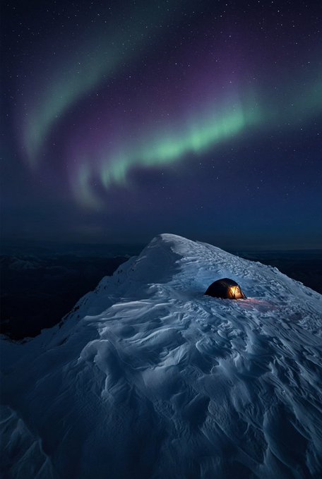 A stunning landscape photograph captures a lone illuminated tent perched on a snowy mountain peak beneath a vibrant display of green and purple aurora borealis lighting up the starry night sky.