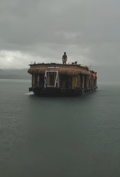 The image features a man standing on top of a boat floating in the water, with another person nearby. The boat appears to be made out of bamboo or straw, adding an interesting element to the scene. The sky above them has some clouds, suggesting that they might be experiencing a cloudy day. In the background, there is a building visible, which could potentially be related to their current situation on the water.