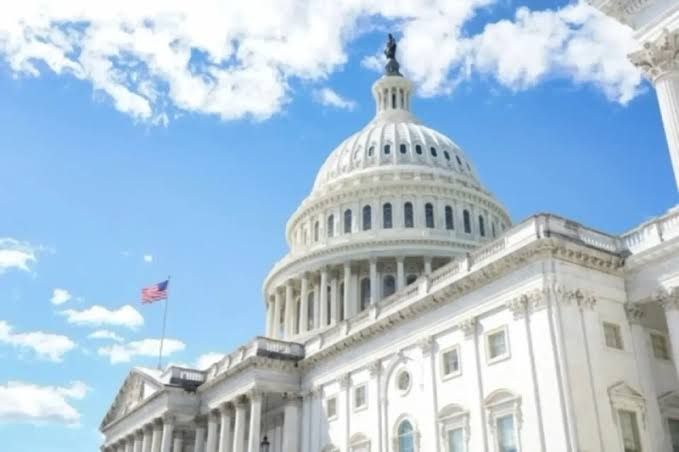 The U.S. Capitol Building is shown against a bright blue sky with scattered clouds, featuring its iconic white dome and an American flag flying.