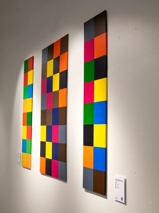 Three tall, rectangular paintings featuring colorful checkerboard patterns in primary colors hang on a white gallery wall.