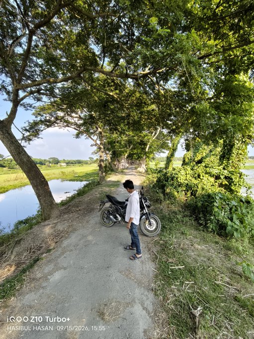 A person in a white shirt and cap stands next to a black motorcycle on a rural dirt path beside a pond, with a watermark reading 'iQOO Z10 Turbo+ HASIBUL HASAN 09/15/2026, 15:55'.