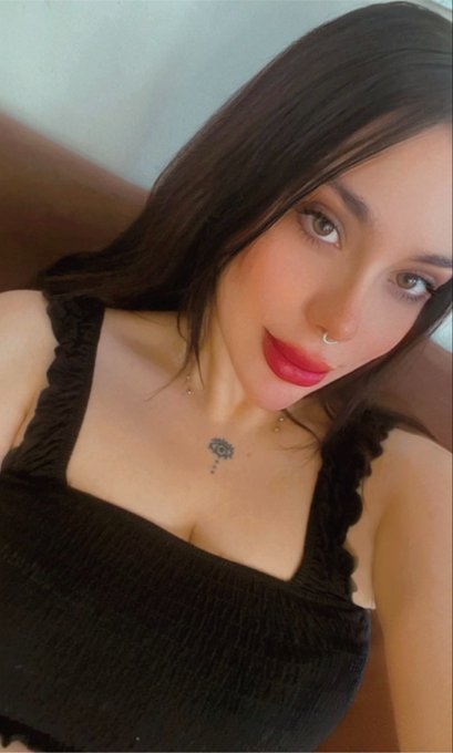 A close-up selfie of a woman with dark hair, red lipstick, and a small chest tattoo, wearing a black top.