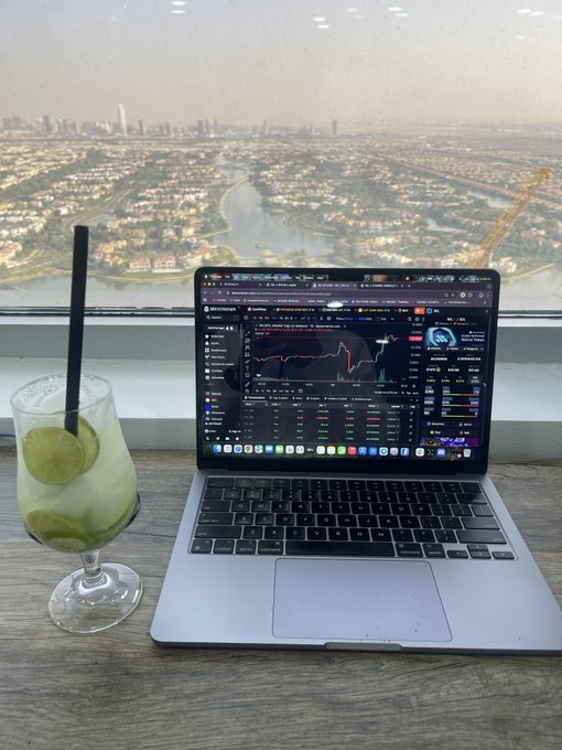 A laptop displaying a cryptocurrency trading chart sits on a wooden desk next to a mojito cocktail, with a sweeping cityscape and river view seen through a large window in the background.