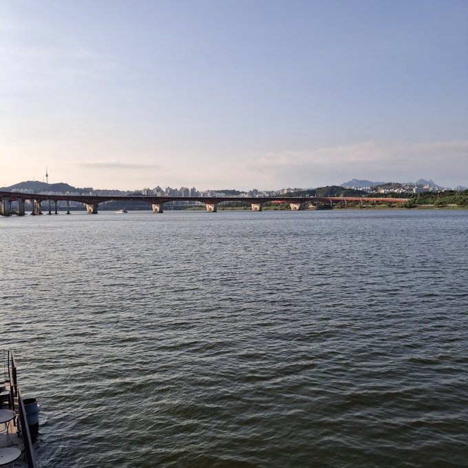 A wide shot of a long bridge spanning a wide river with a city skyline and mountains visible in the background under a hazy sky.