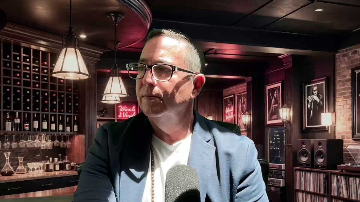 The image features a man wearing glasses, standing in front of a bar with wine bottles displayed behind him. He appears to be talking into a microphone or recording device, possibly discussing something related to the wine selection at the bar. The background includes a brick wall and several lights hanging from the ceiling, creating an atmosphere for socializing and enjoying drinks.