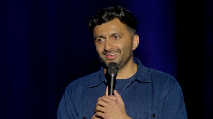 A man with dark hair, wearing a blue denim shirt, holds a microphone and smiles while performing on stage against a dark blue curtain backdrop—likely comedian Nimesh Patel, whose special "BUY THE DIP" is now streaming on Netflix, as referenced in the caption.